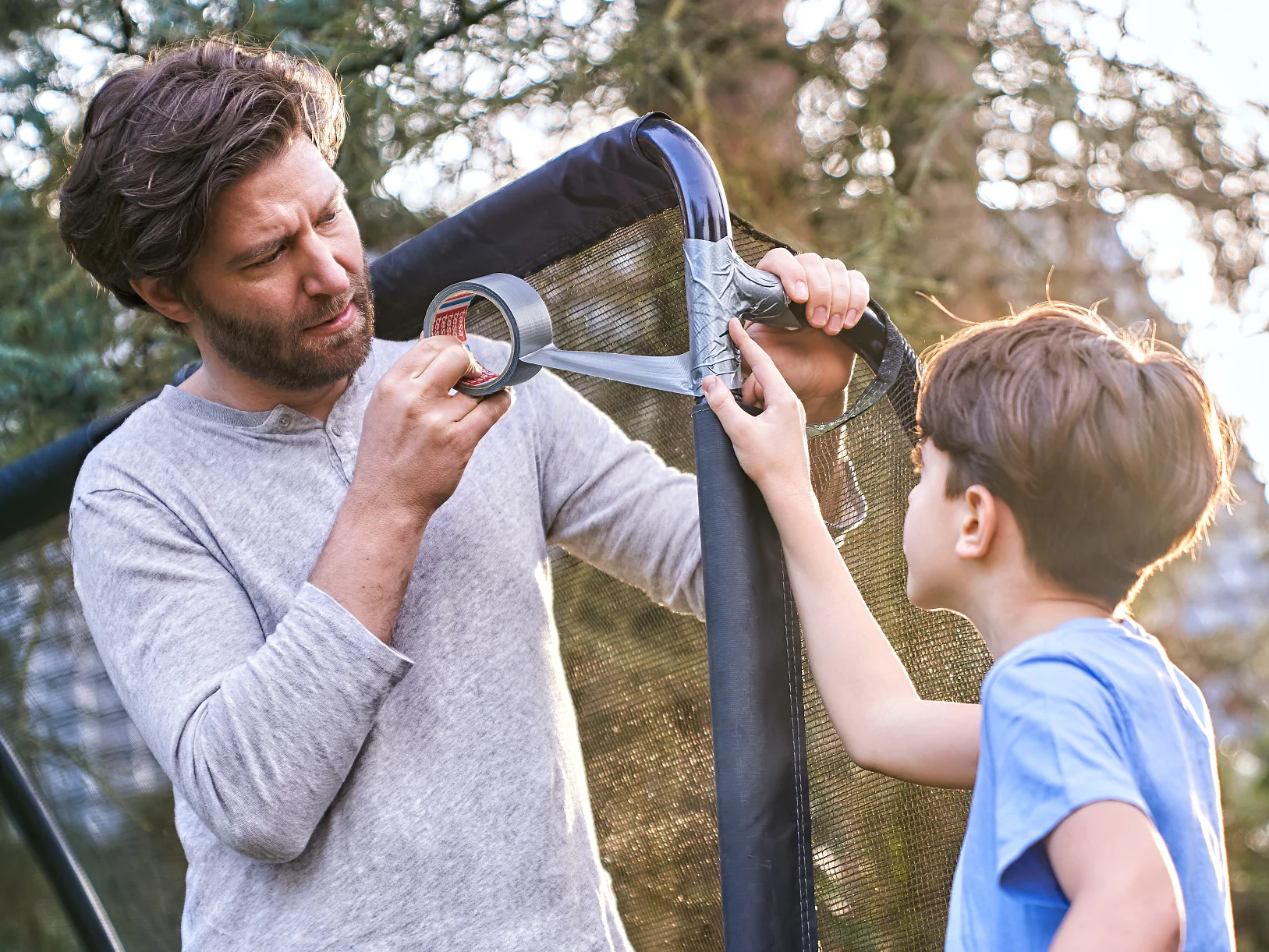 A man is using tesa tape to repair a net structure outdoors while a boy holding a football watches. They are standing near trees, with the sun shining in the background. The scene suggests a collaborative effort. (This text has been generated by AI)