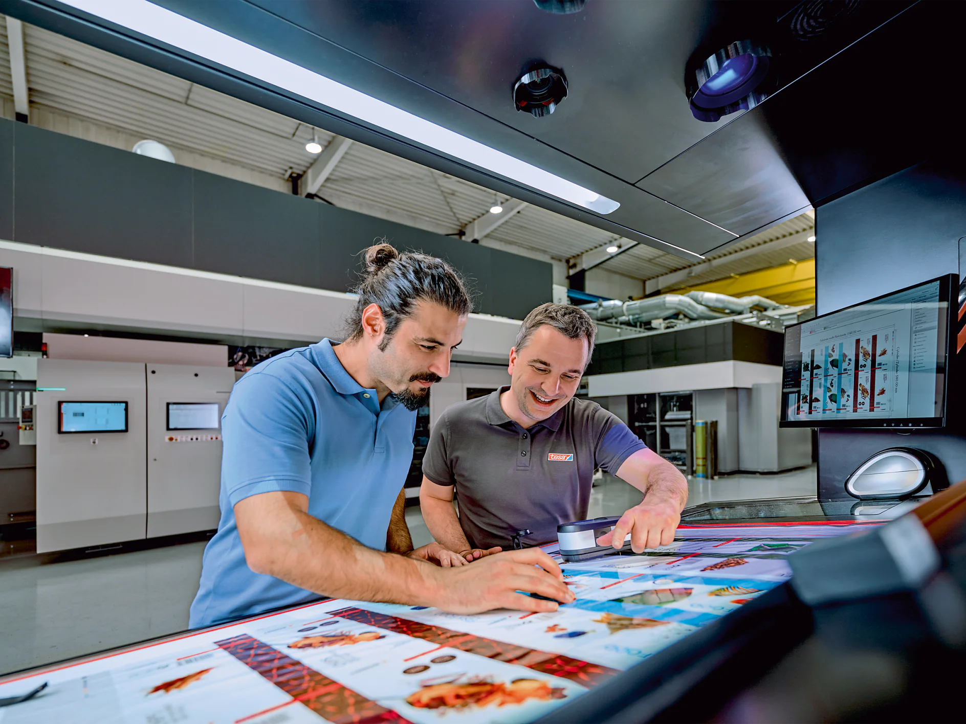 Two men stand at a large control panel reviewing printed sheets in a modern industrial printing facility. One man uses a magnifying tool while both examine the color quality and application of tesa tape. Monitors and equipment are visible in the background. (This text has been generated by AI)