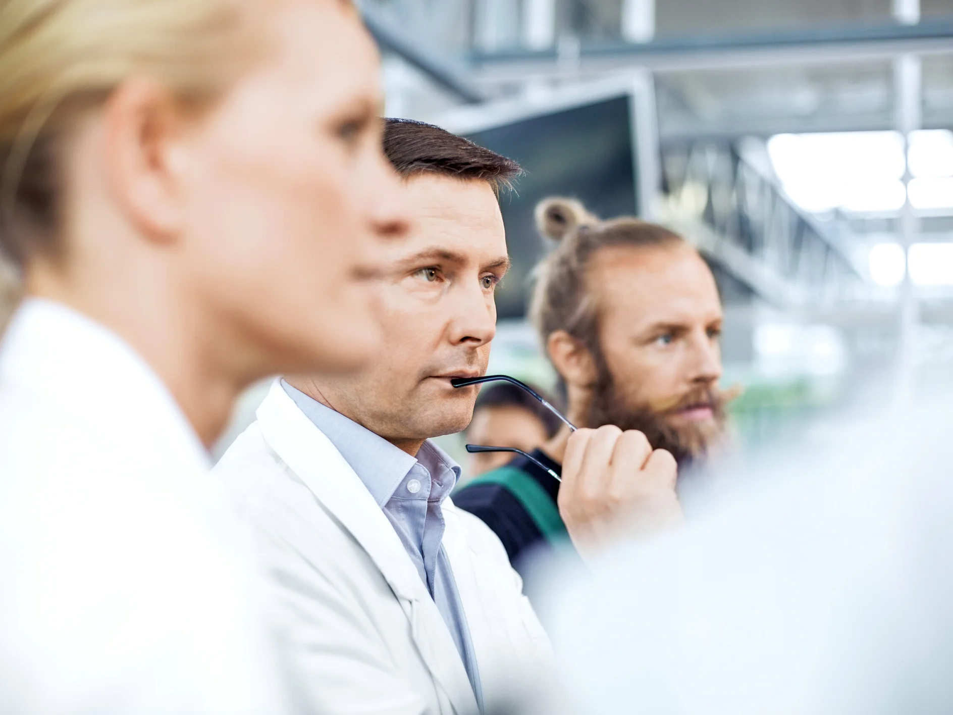 Three people in professional attire are shown in close-up, sitting indoors. The man in the center holds eyeglasses near his chin, looking thoughtful, while the others look in the same direction. A roll of tesa tape is visible nearby. The background is blurred. (This text has been generated by AI)