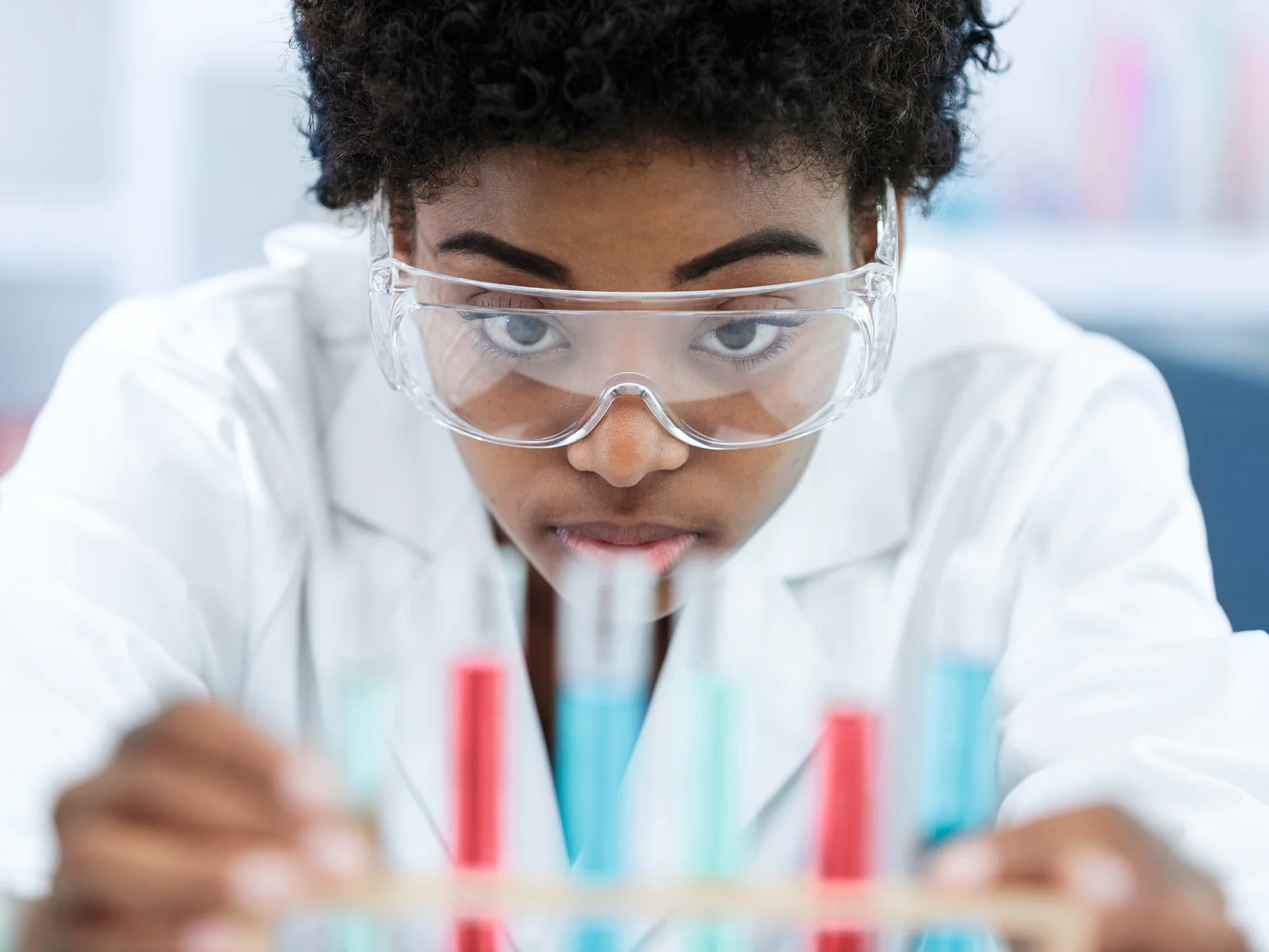 A person wearing safety goggles and a white lab coat focuses intently on several test tubes filled with colorful liquids, with tesa tape visible in the laboratory or scientific setting. (This text has been generated by AI)
