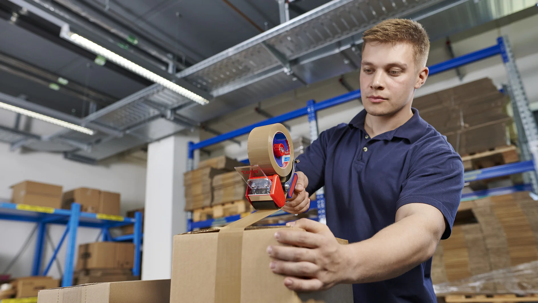 a person in a blue shirt is sealing a cardboard box with packing tape in a warehouse filled with stacked boxes and shelves.
