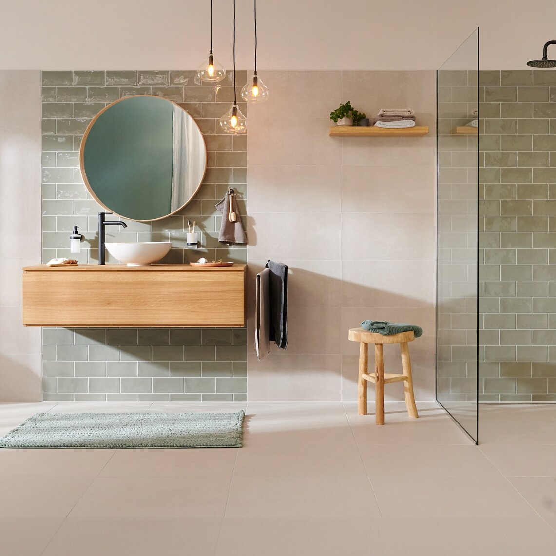 Modern bathroom with beige tiles and green tiled shower area. Features include a wall-mounted wooden vanity with a round mirror, a white toilet, a glass shower partition secured with tesa tape, hanging lights, and a wooden stool. A towel hangs near the shower using tesa. The bathroom is now completely tidy, as also a shower basket is now holding the shower soaps. (This text has been generated by AI)