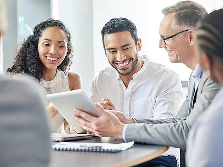 Four people are sitting at a table, looking at a tablet, smiling and engaged in discussion. Notebooks, papers, and tesa tape are on the table. The setting appears to be a modern office or meeting room. (This text has been generated by AI)