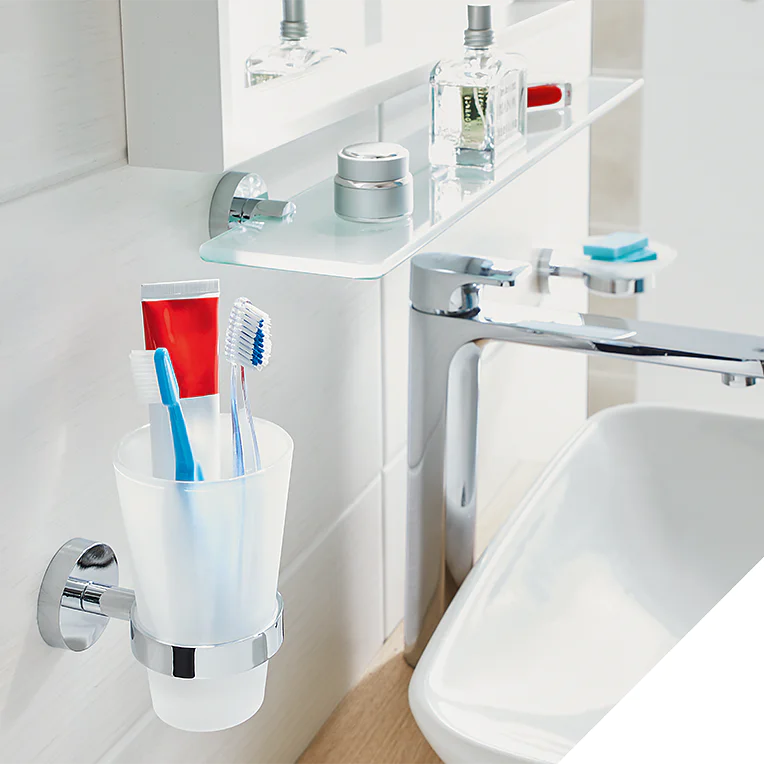 A bathroom sink area features a white cup holding toothbrushes and toothpaste mounted on the wall using tesa tape. Above, a shelf holds toiletries. A silver faucet is attached to the sink. On the right, a silver wall hook is shown. (This text has been generated by AI)