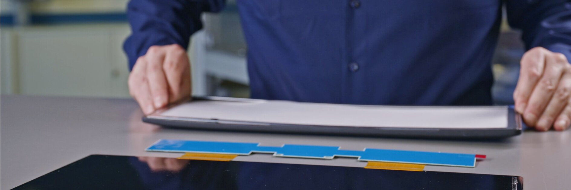 person in blue shirt holding rolled-up paper near tablet and colorful pens on white desk in indoor workspace