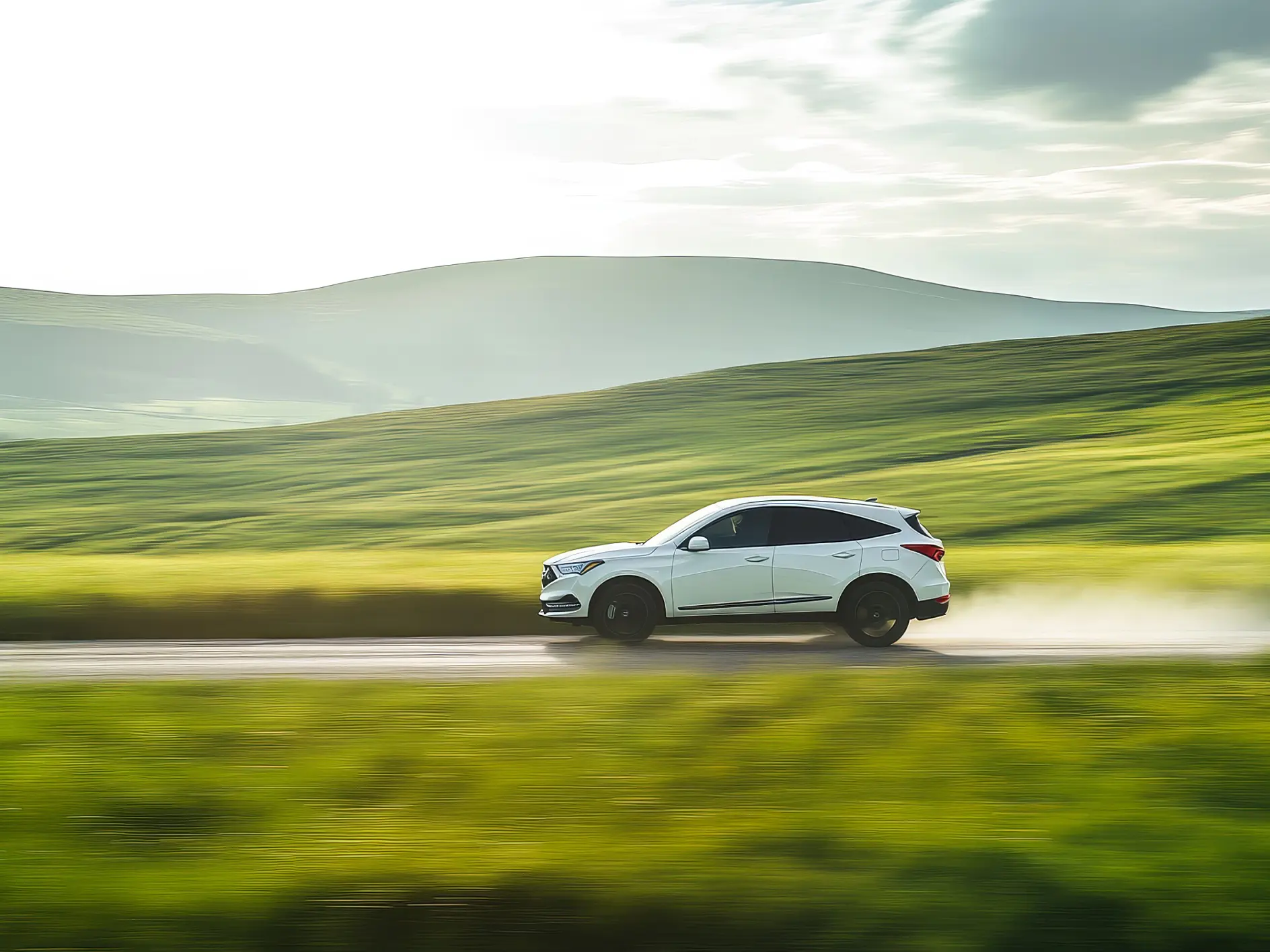 A white SUV speeds along a paved road among green hills, with blurred grass in front and hills behind beneath a partly cloudy sky.
