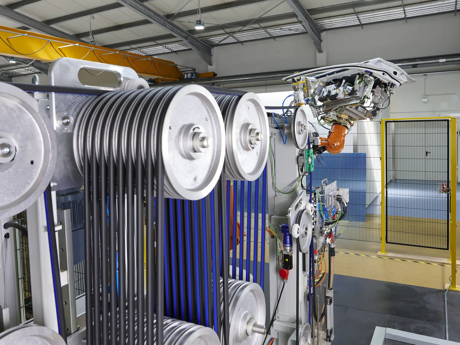 A close-up of industrial machinery with multiple pulleys and blue tesa tape belts, a robotic arm in the background, and a fenced area nearby.