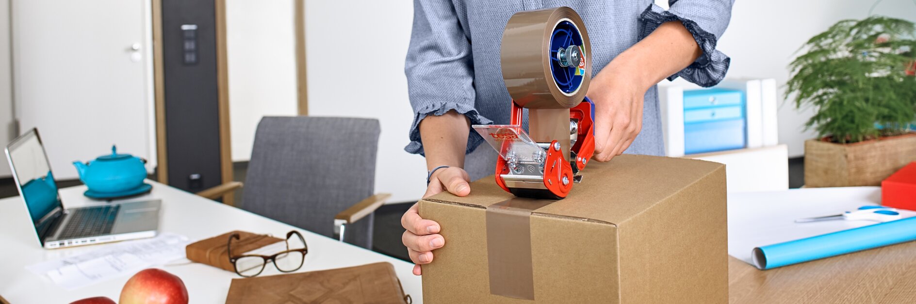 Person sealing a cardboard box with brown packing tape using a tape dispenser, on a desk with office supplies and fruit in the background.