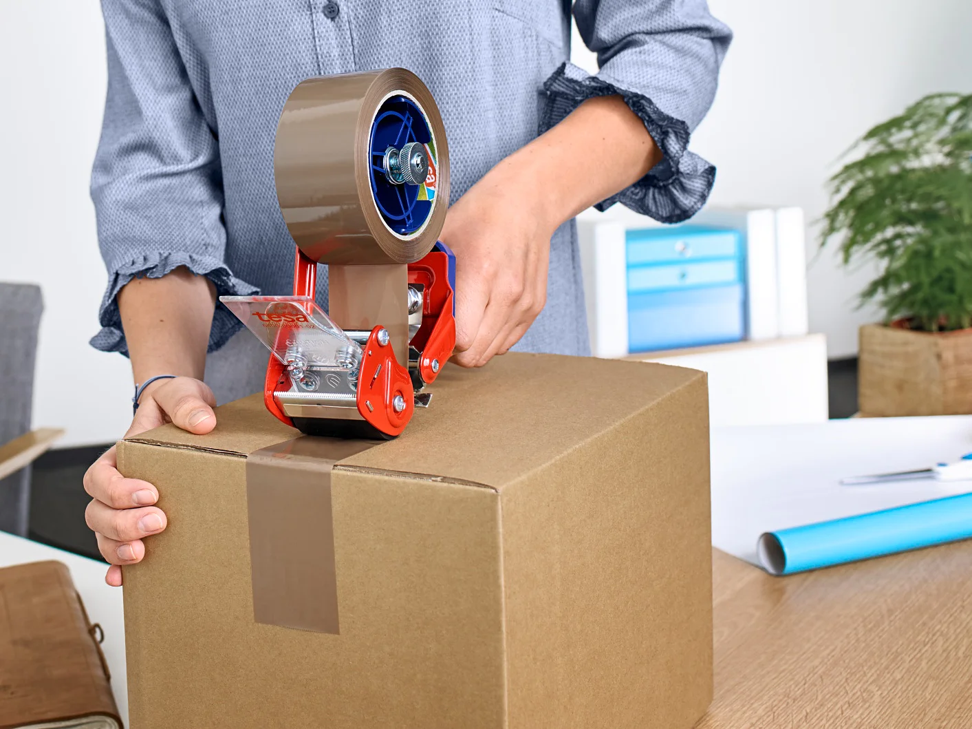 Person sealing a cardboard box with brown packing tape using a tape dispenser, on a desk with office supplies and fruit in the background.