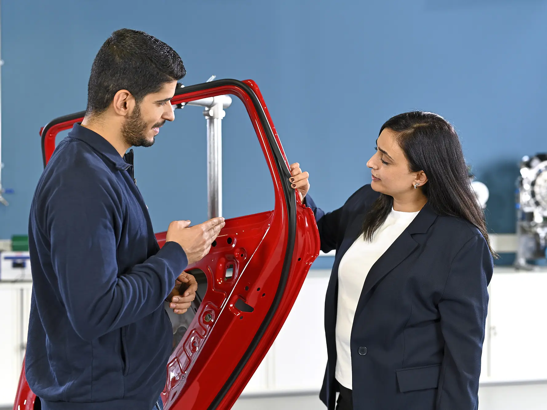Two people in a workshop discuss a vertical red car door, with one gesturing and the other holding it; tesa tape is visible nearby.