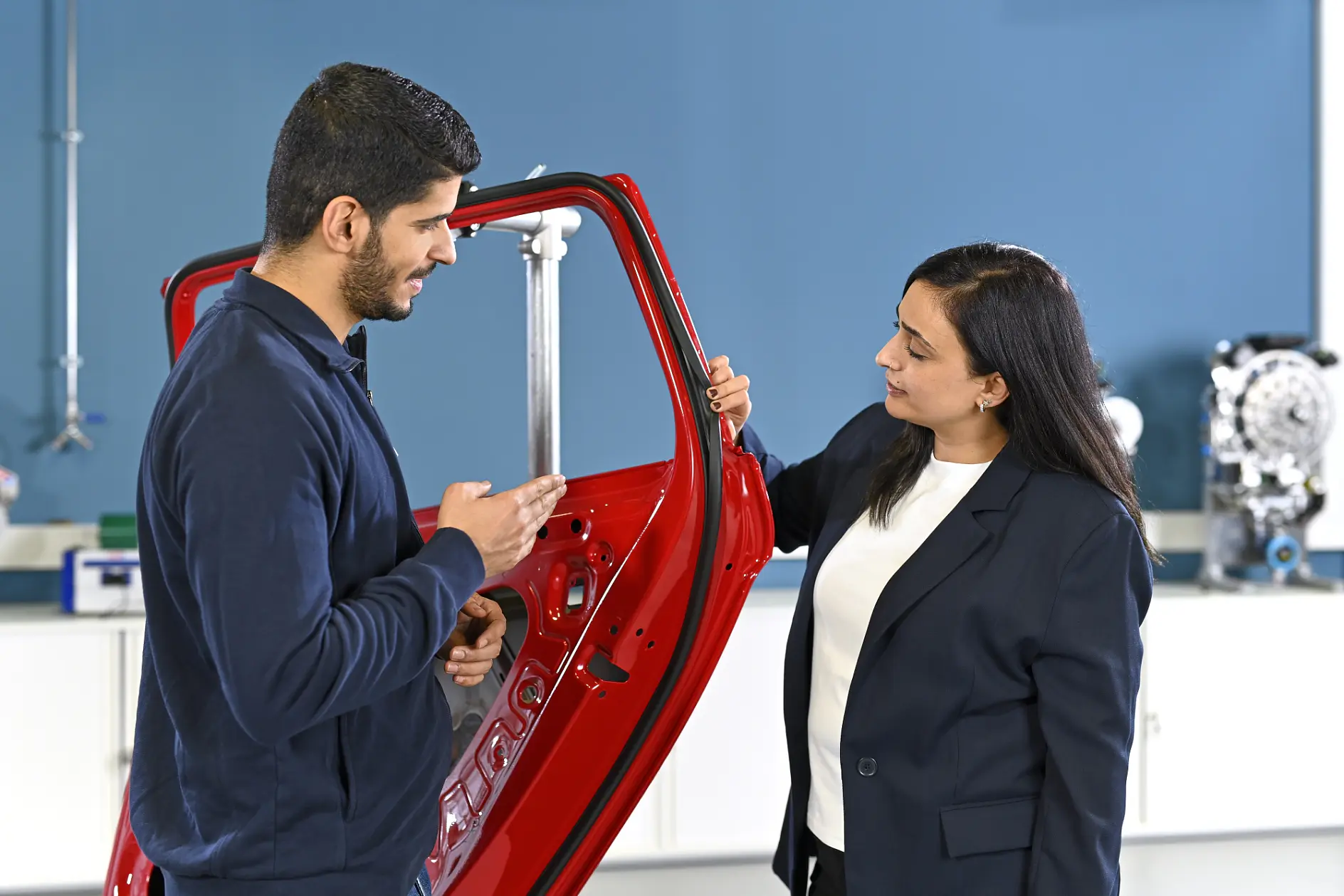 Two people in a workshop discuss a vertical red car door, with one gesturing and the other holding it; tesa tape is visible nearby.