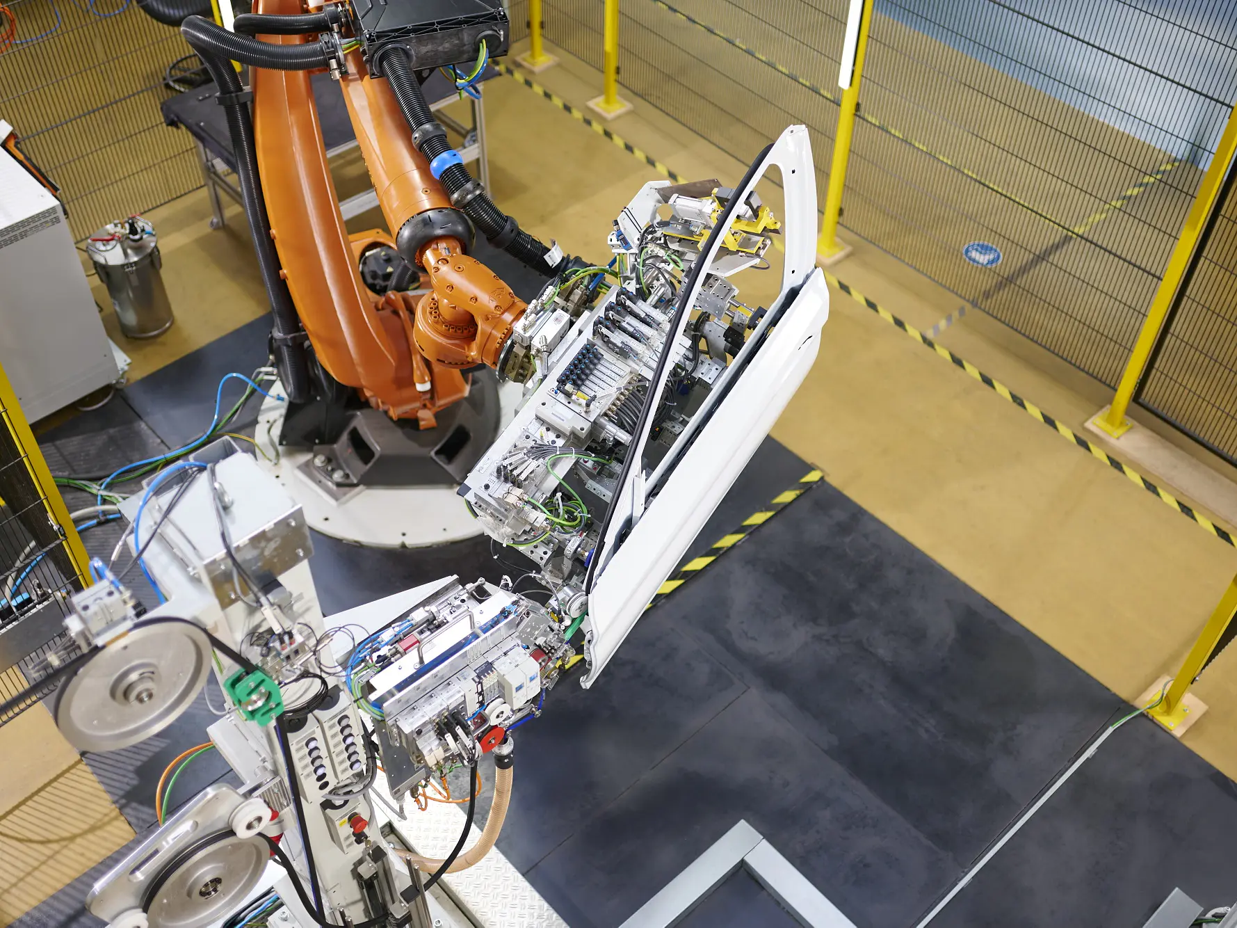 A robotic arm installs a car door with tesa tape onto a vehicle frame in an industrial facility, surrounded by safety barriers and yellow railings.