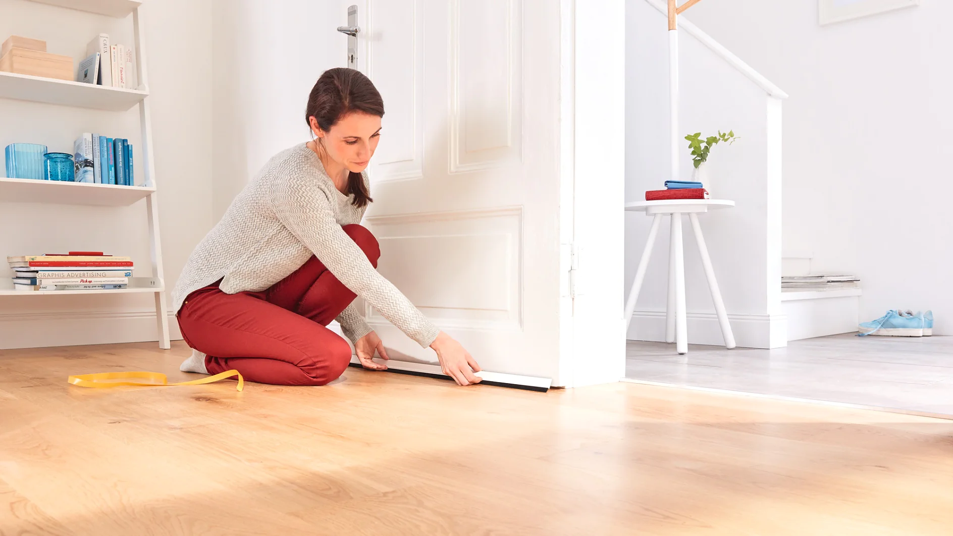 A person is kneeling on a wooden floor, fitting a draught excluder to the bottom of a white door. Next to them, a Tesa tape measure is lying on the floor. In the room, there is a staircase, a bookshelf, and a small table with a plant. (This text has been generated by AI)