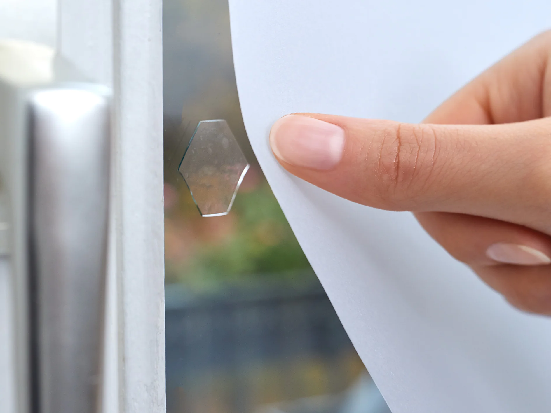 Hand lifting a frosted window film to show a clear hexagonal adhesive tab on the glass.