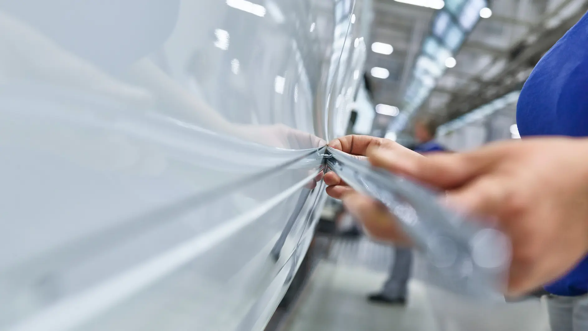 tesa tape being applied to a white vehicle in a factory setting, focusing on hands smoothing the tape along the surface (This text has been generated by AI)