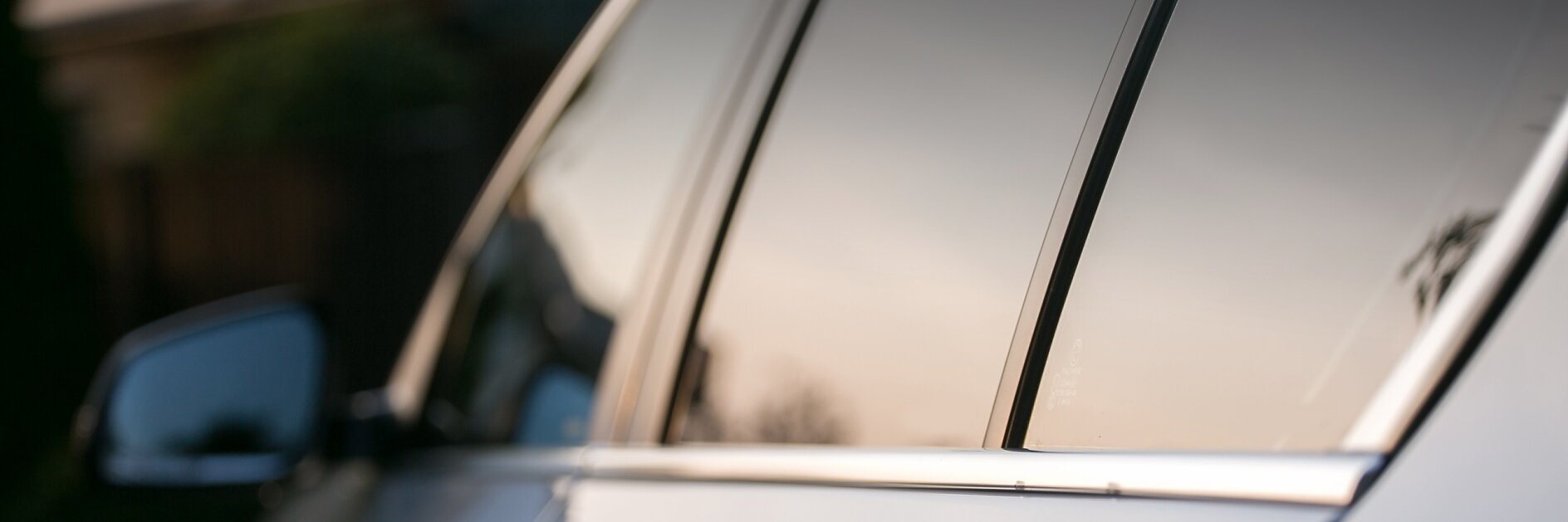 Side view of a silver car reflecting the soft light of a setting sun. The focus is on the side windows and door handle, with blurred greenery in the background.