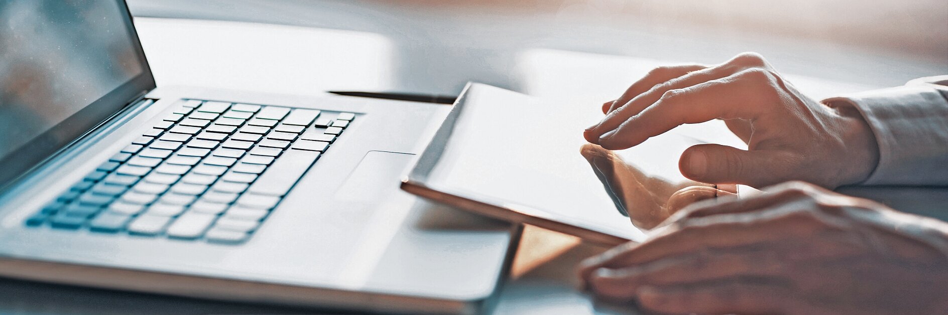hands writing in a notebook beside an open laptop on a desk in a bright indoor workspace