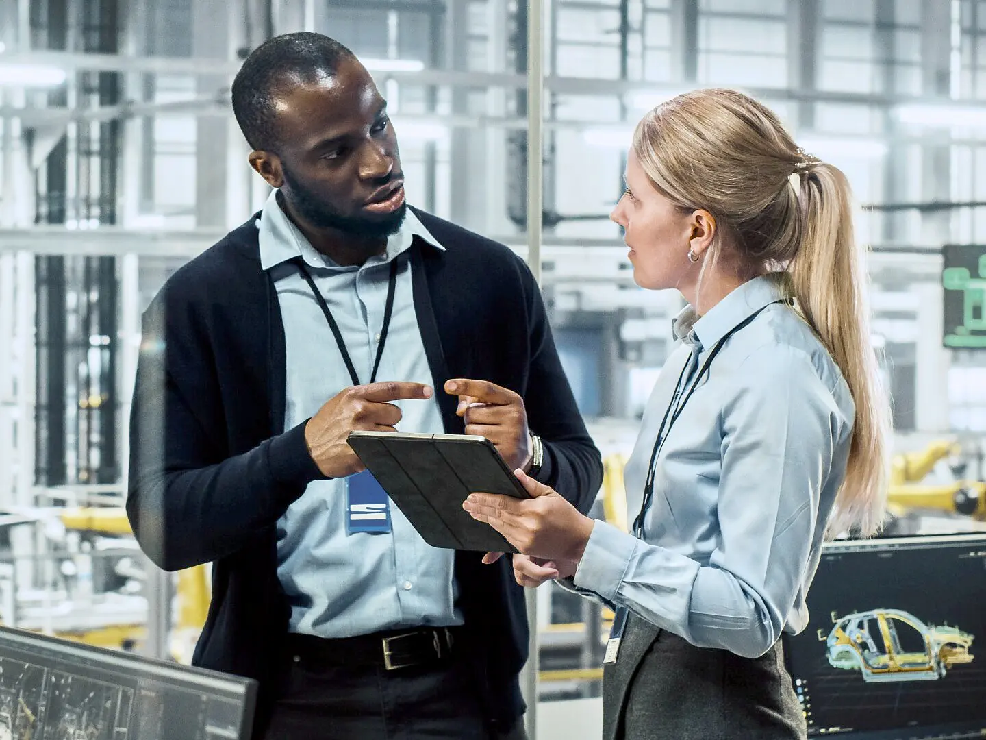 Two professionals in business attire discuss information on a tablet in an industrial office; tesa tape, machinery, and monitors are visible. (This text has been generated by AI)