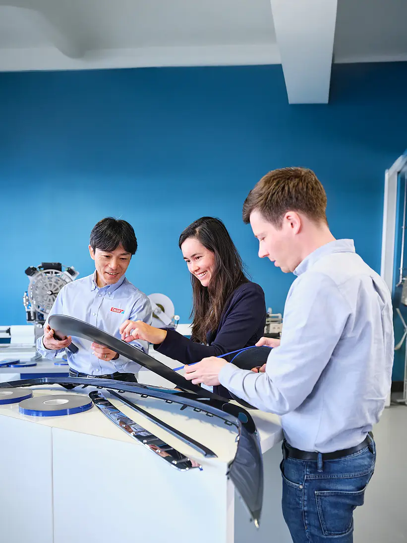 Three people stand at a table examining automotive parts and tesa tape in a bright, modern workspace with blue walls and equipment. (This text has been generated by AI)