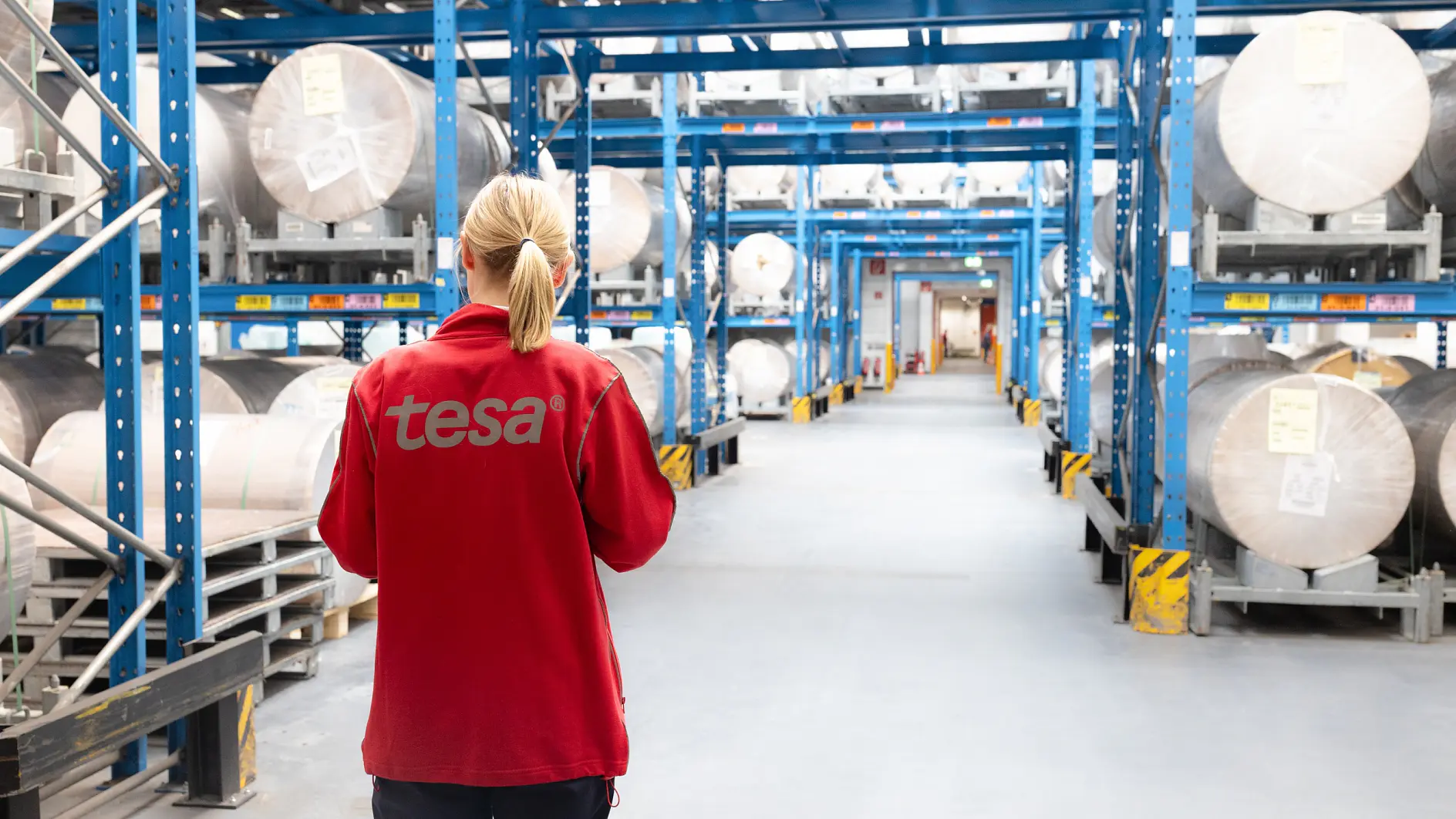 A person in a red jacket with tesa on the back stands in a warehouse aisle, surrounded by large rolls of tesa tape on blue shelves. (This text has been generated by AI)