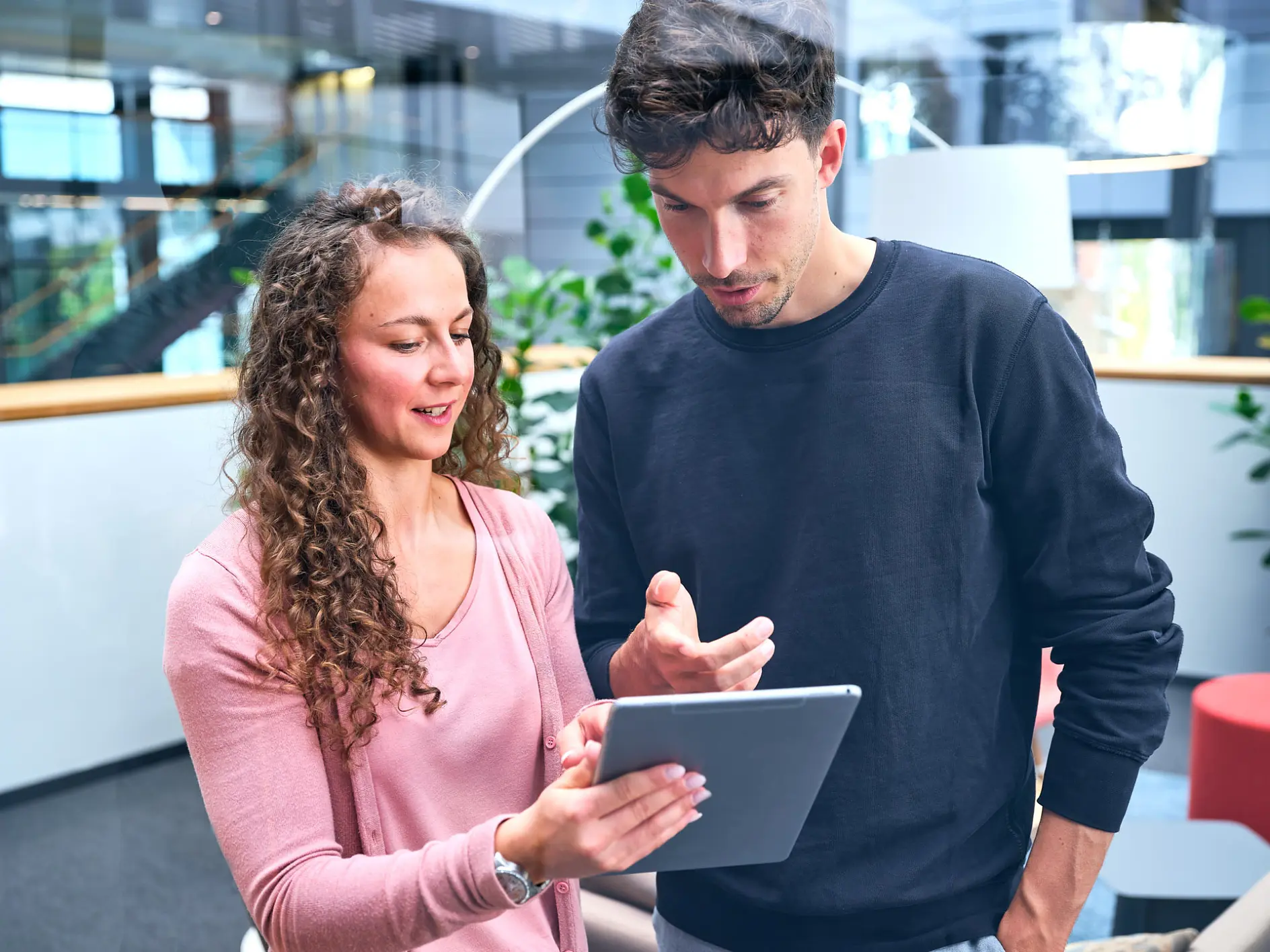 A woman and a man are standing in a modern office space. The woman is holding a tablet and gesturing towards it, while the man looks at the screen attentively. They are engaged in discussion, with plants and office furniture in the background. (This text has been generated by AI)