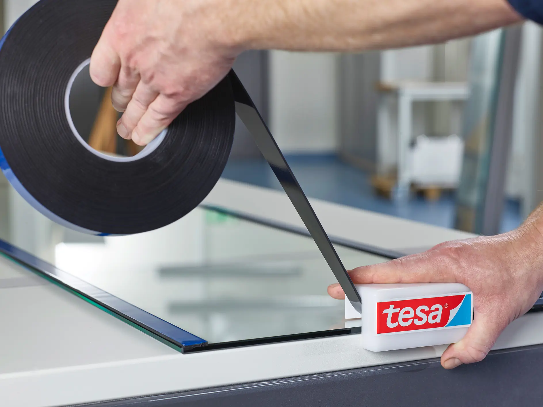hands applying black tesa tape to glass panel edge indoors, focusing on precise alignment and smooth application