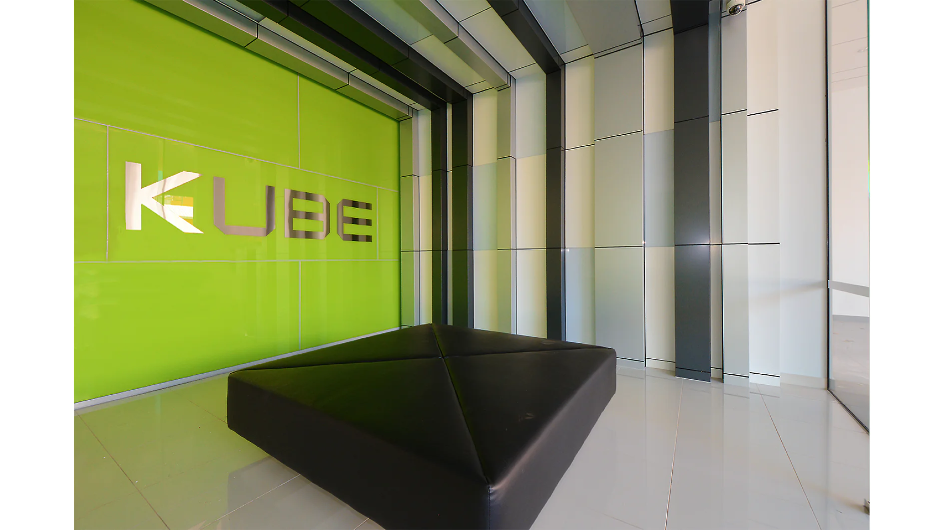Interior room with a green sign on the wall reading KUBE in stylized letters. The walls have a metallic striped design, and there is a large triangular black cushion on the shiny tiled floor, all assembled and accentuated using tesa tape. (This text has been generated by AI)