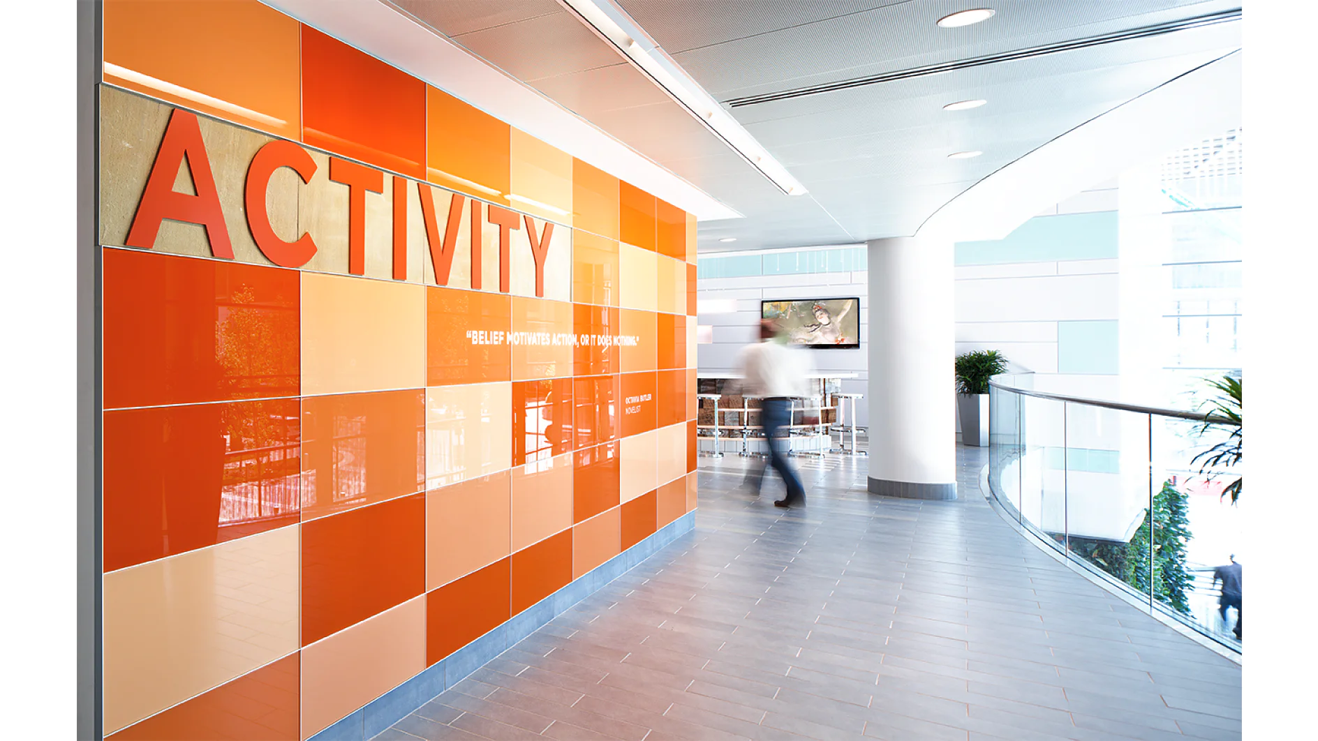 A hallway with a glossy tile wall in shades of orange and red, spelling out ACTIVITY in large letters using tesa tape. A person walks past, and a seating area with tables and chairs is visible in the background. (This text has been generated by AI)