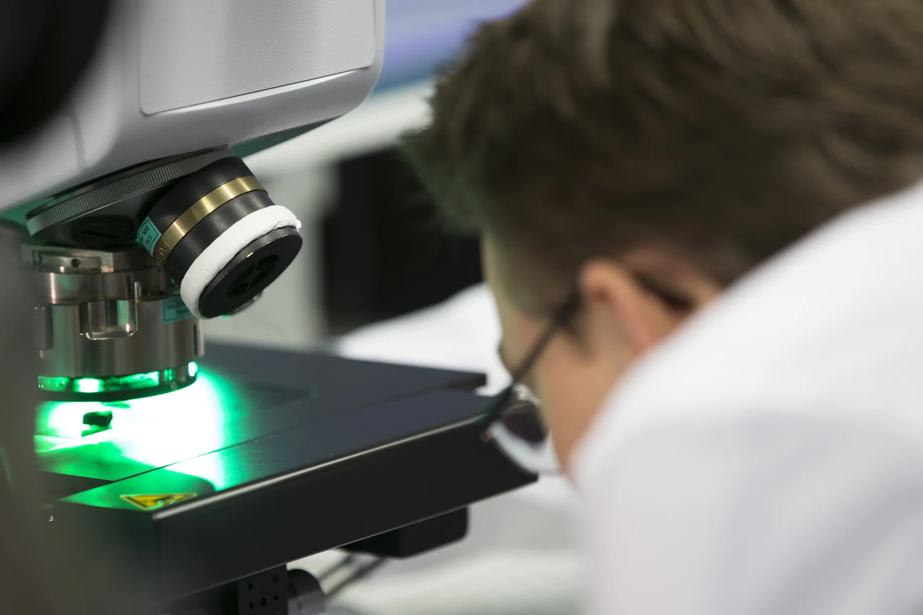 A person wearing glasses closely observes a sample under a microscope emitting green light. The focus is on the equipment, with blurred background elements suggesting a laboratory setting, and nearby, tesa tape is ready for any necessary applications. (This text has been generated by AI)