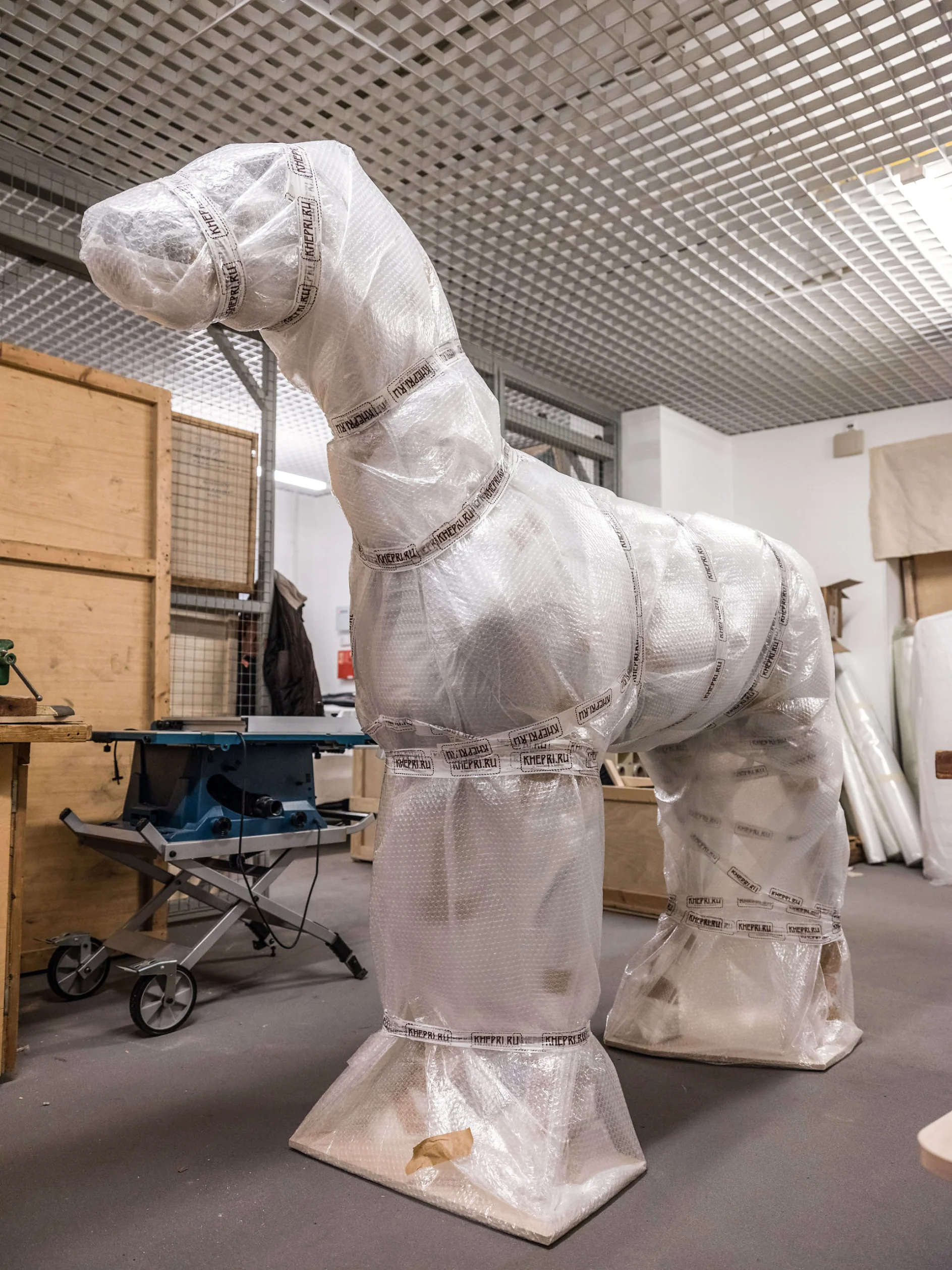 A large sculpture wrapped in bubble wrap and tesa tape resembles a horse with an elongated neck. The scene is set in an indoor workspace with wooden crates and various tools in the background. (This text has been generated by AI)