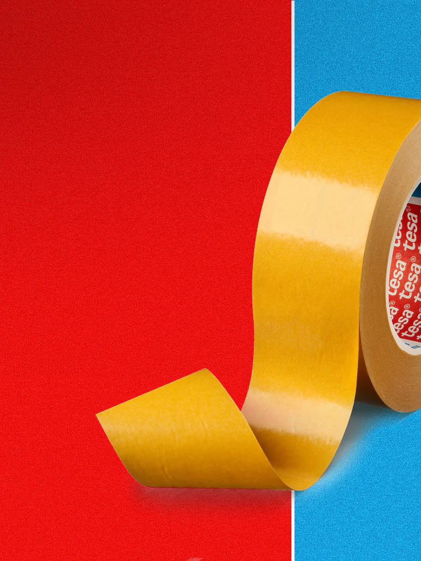 A roll of yellow tesa tape stands upright against a red and blue split background. The loose end of the tesa tape curls outward. The inside of the roll has a red label with white text. (This text has been generated by AI)