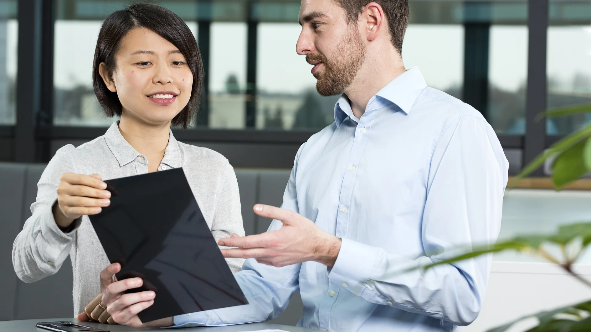 Two people are discussing something on a tablet in an office setting. The woman is holding the tablet, while the man gestures toward it. A document with graphs is on the table secured with tesa tape. They are seated near a window with a view outside. (This text has been generated by AI)