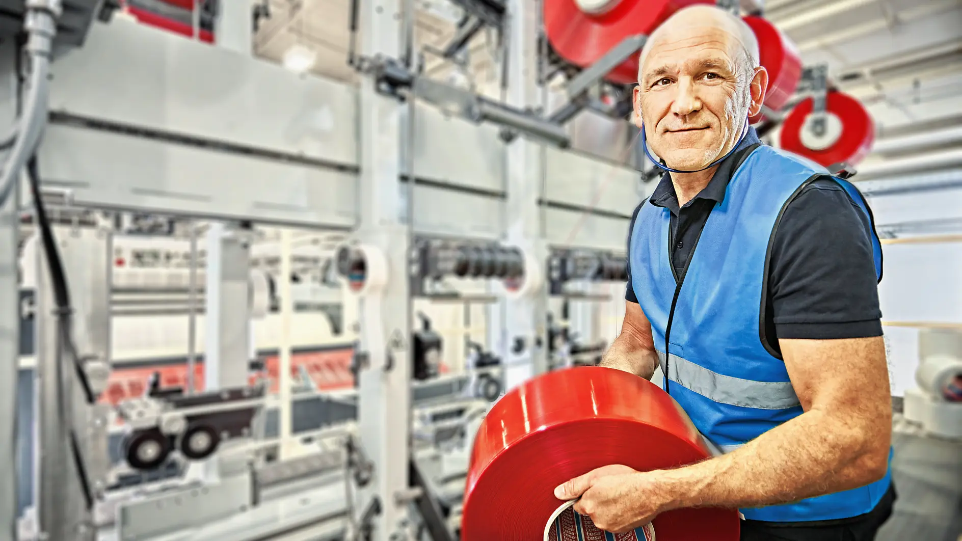 A man in a blue safety vest holds a large red roll of tesa tape in a factory setting. Behind him are industrial machines with more red rolls of tesa tape. The environment is well-lit and industrial.