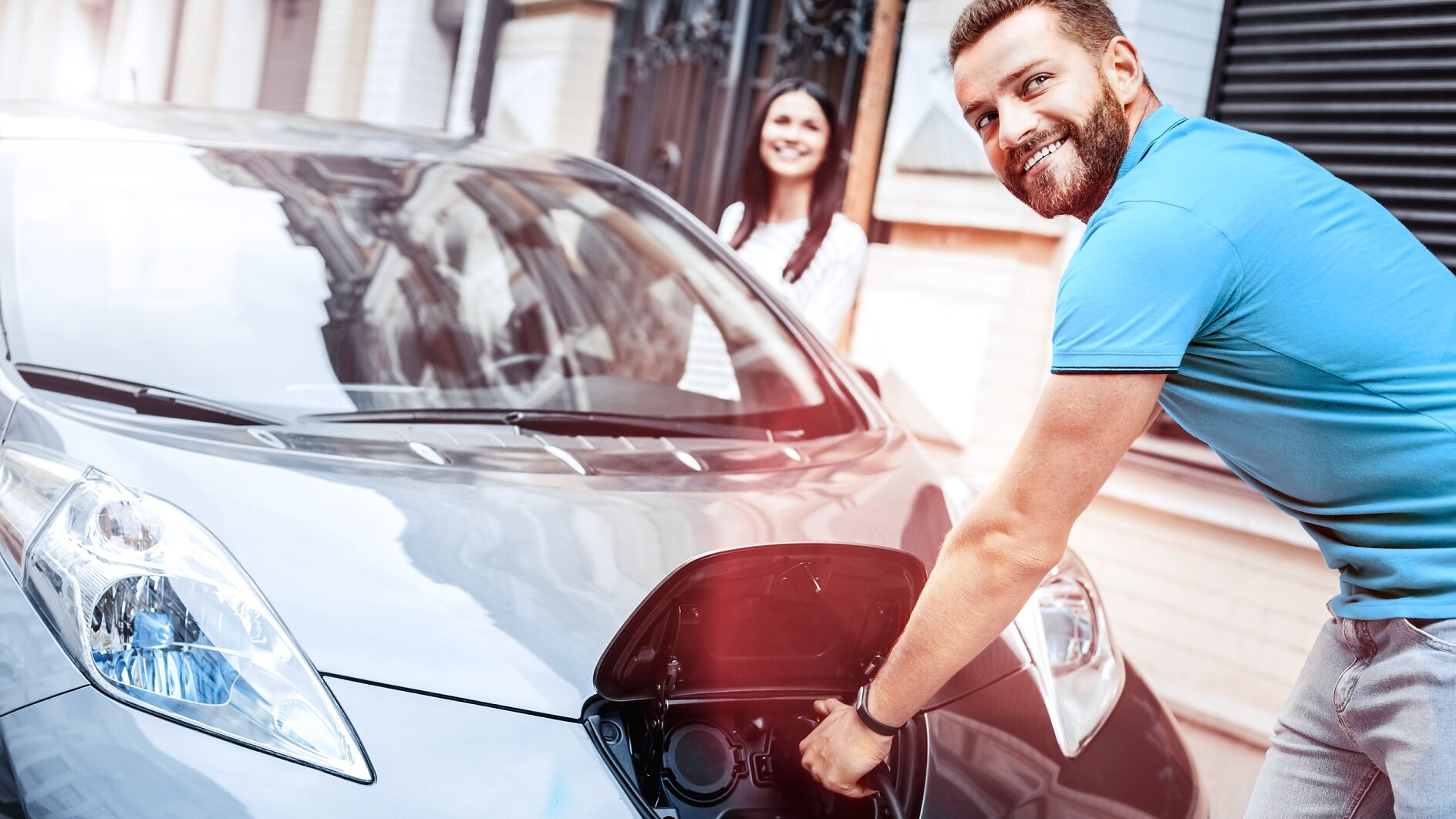 A man is plugging a charging cable into an electric car on a city street using tesa tape to secure the cable. The car is silver, and its front charging port is open. A woman stands nearby, smiling at the man as he uses tesa tape. Theyre in front of a building with a brick facade. (This text has been generated by AI)