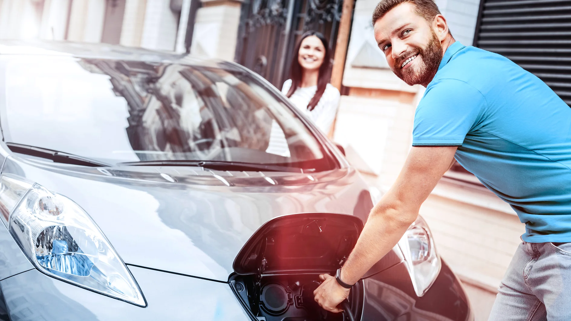 A man is plugging a charging cable into an electric car on a city street using tesa tape to secure the cable. The car is silver, and its front charging port is open. A woman stands nearby, smiling at the man as he uses tesa tape. Theyre in front of a building with a brick facade. (This text has been generated by AI)