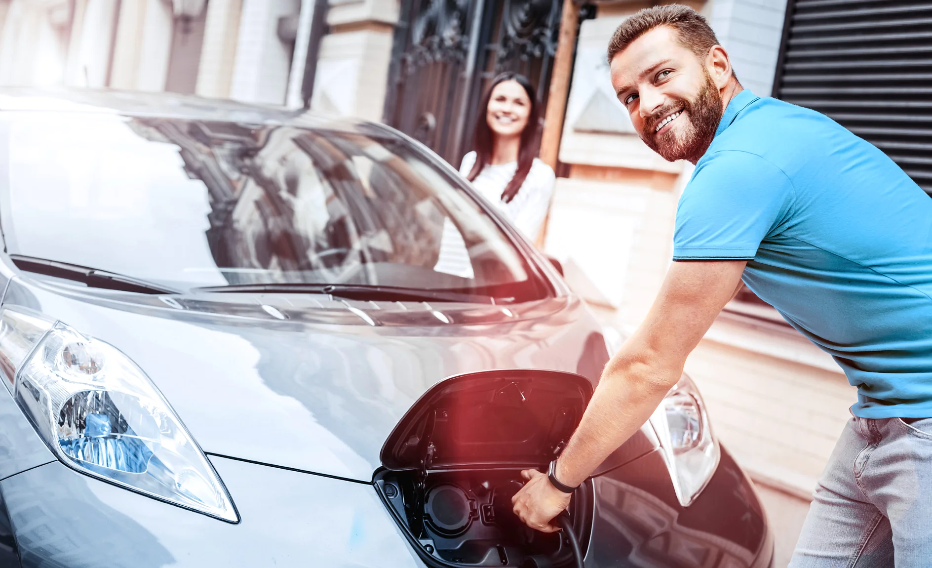 A man is plugging a charging cable into an electric car on a city street using tesa tape to secure the cable. The car is silver, and its front charging port is open. A woman stands nearby, smiling at the man as he uses tesa tape. Theyre in front of a building with a brick facade. (This text has been generated by AI)