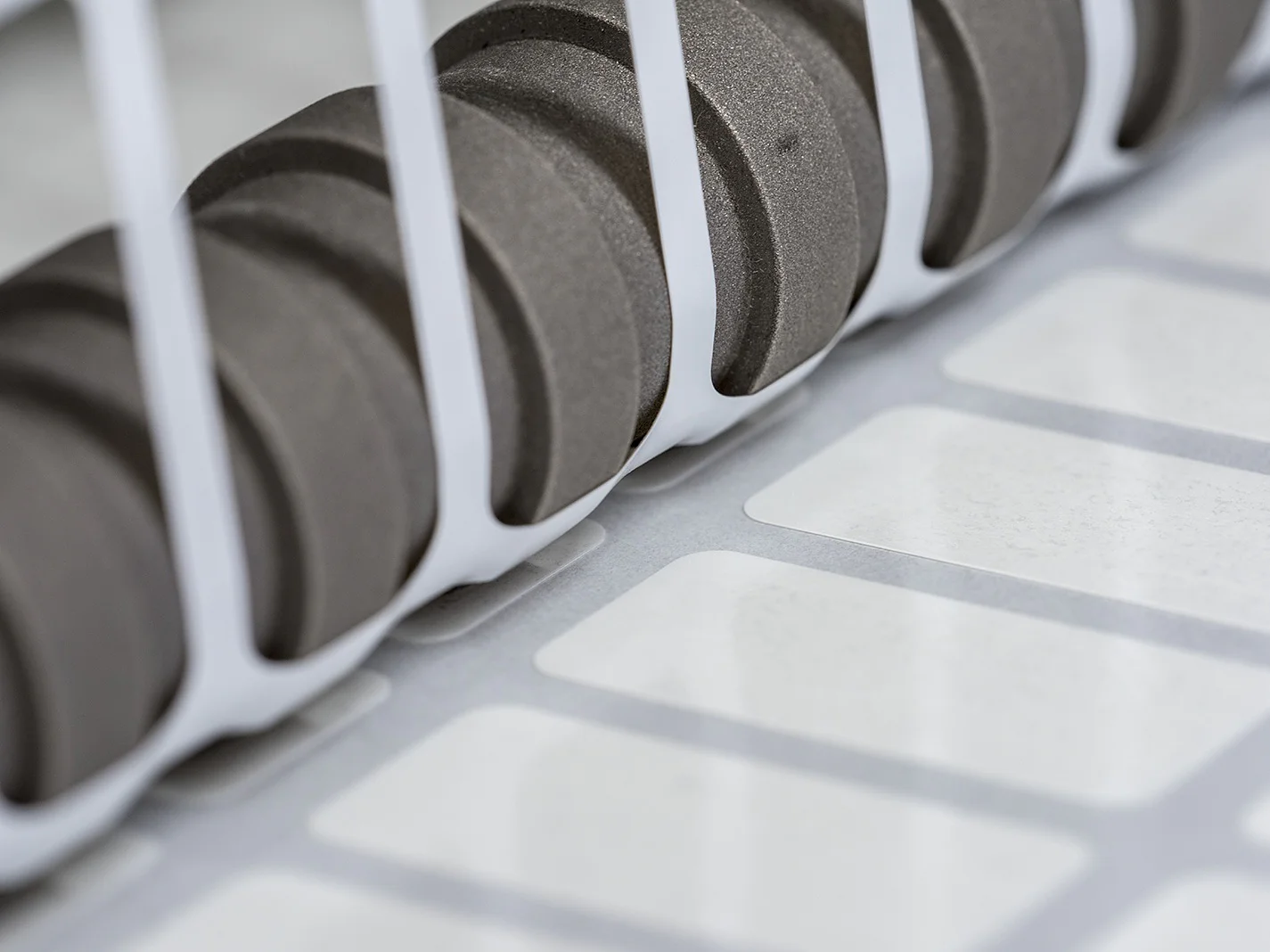 Close-up of a set of black gears positioned next to a row of white labels running along a conveyor system, partially enclosed by white vertical bars. The scene suggests an industrial setting, likely related to printing or labeling using tesa tape. (This text has been generated by AI)