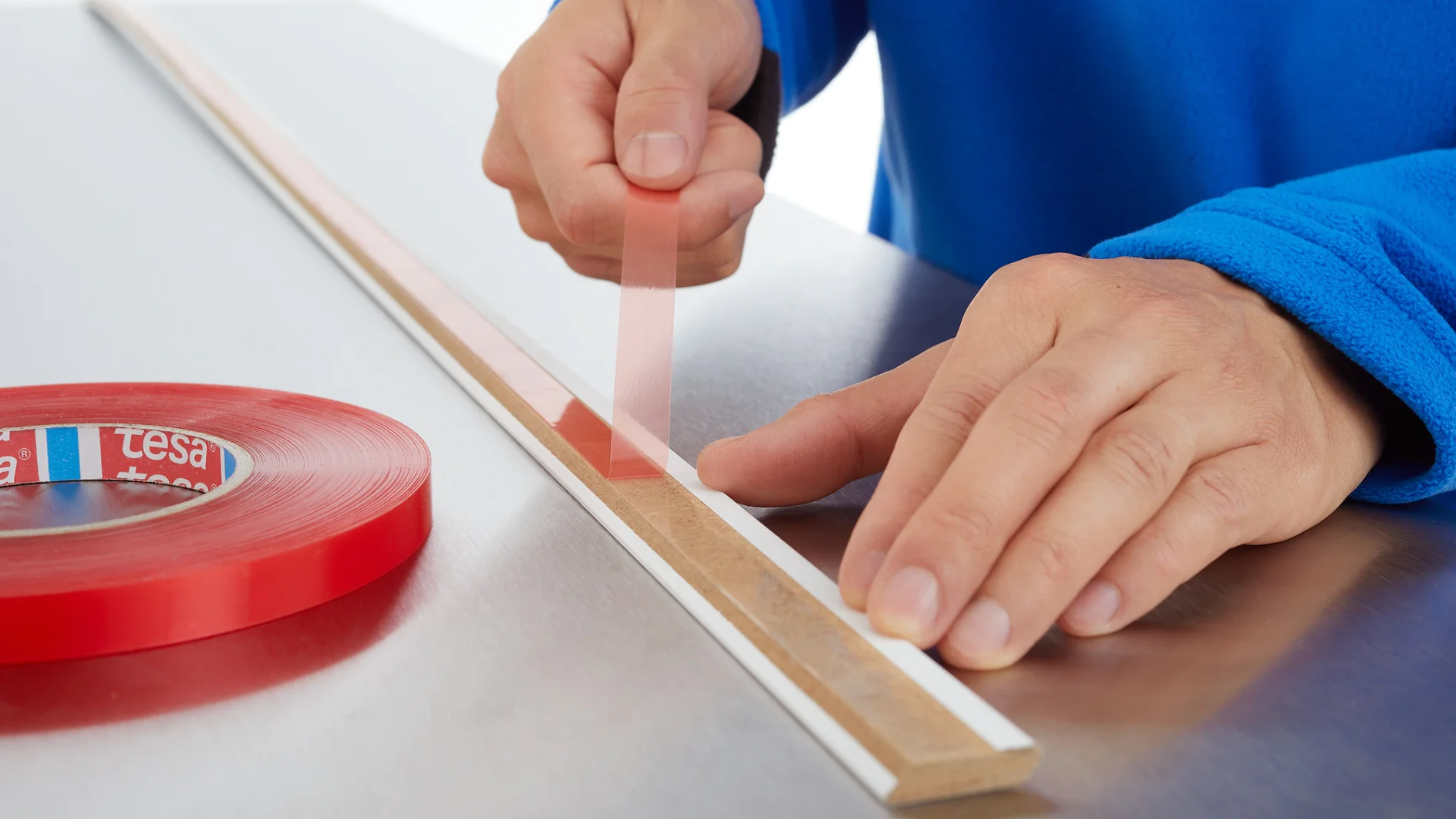 A person wearing a blue sleeve applies red double-sided adhesive tesa tape to a long, thin white strip on a metal surface. A red roll of tesa tape is positioned beside them. (This text has been generated by AI)