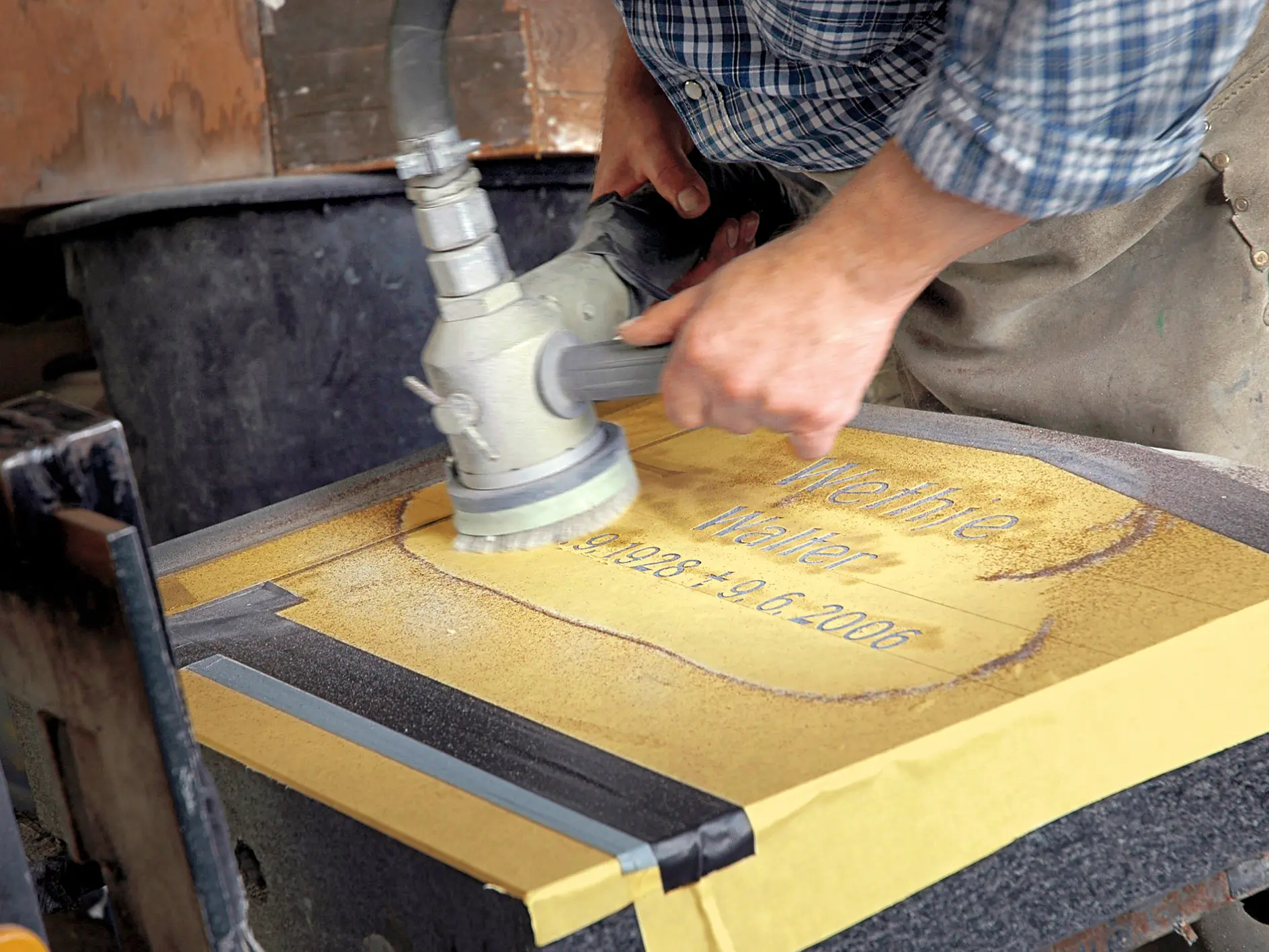 A person using a power tool to sand or polish a stone surface with the words Meinhte Walter, 9.1.1924 - 9.6.2006 visible on it. The stone is secured around the edges with tesa tape, and the person is wearing a plaid shirt and apron.