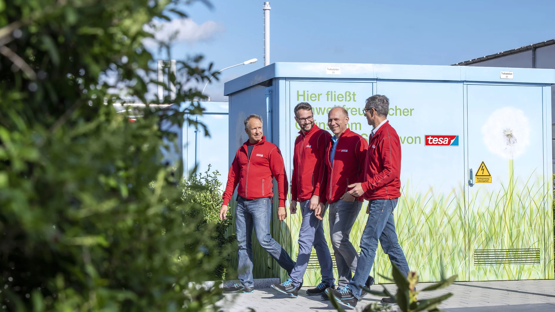 twHH_Environment 5 300dpi Four men in red jackets walk together on a paved path. Behind them is a blue utility structure featuring grass and dandelion artwork. The background includes green foliage under a blue sky. A yellow caution sign is visible on the structure. (This text has been generated by AI)