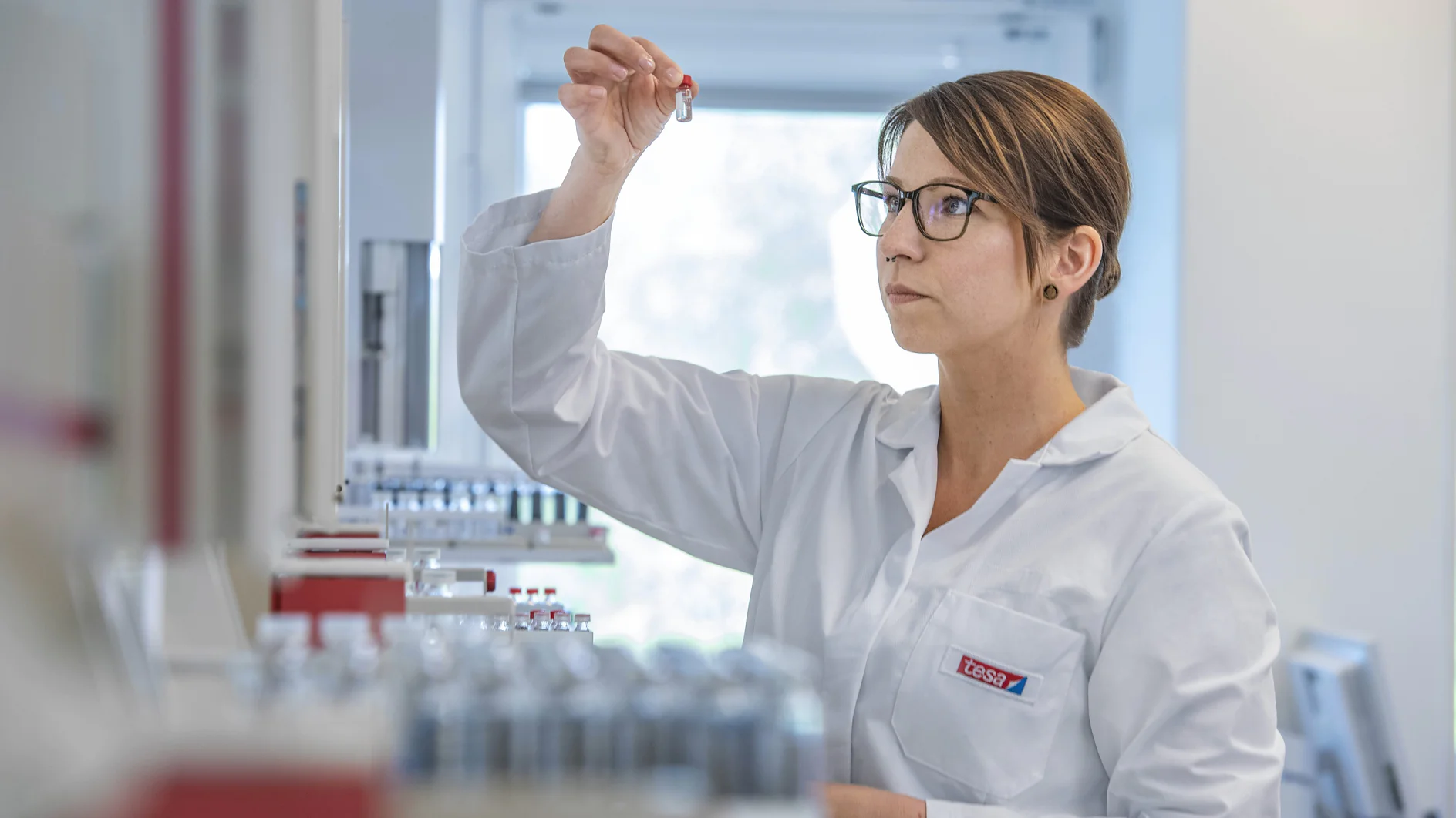 twHH_RandD 3 300dpi A person wearing a white lab coat and glasses is examining a small vial in a laboratory setting. Shelves in the background hold several similar vials and various pieces of laboratory equipment, including rolls of tesa tape. The overall atmosphere is professional and focused. (This text has been generated by AI)