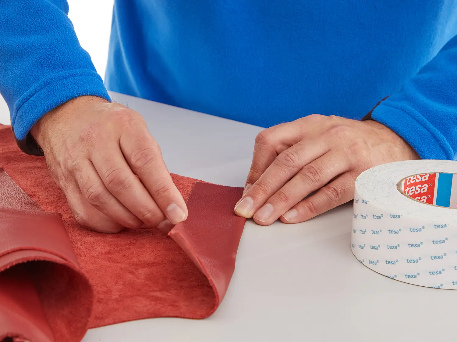 A person wearing a blue fleece folds a piece of red leather-like fabric on a white table, carefully holding the edge. A roll of white tesa tape with red and blue labels is placed nearby, showcasing applications of tissue tapes and tissue tape double sided solutions.