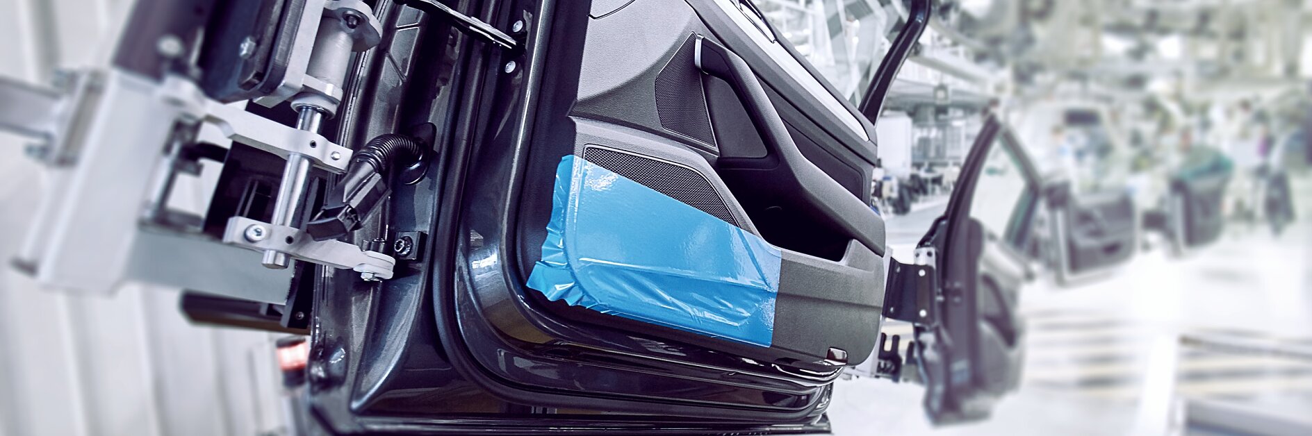 A car door is mounted on an assembly line in an industrial setting. The interior side of the door is visible, partially covered with blue protective tesa surface protection tape. The background shows more equipment and blurred parts of the assembly line.