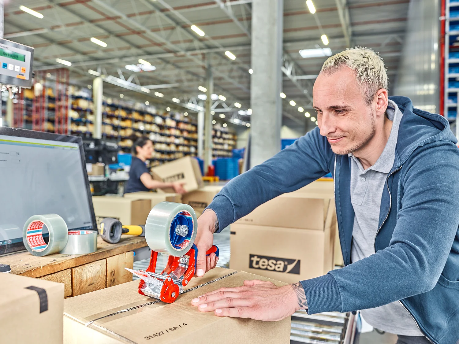TESA-60412_MM_24655_final_72dpi A person is sealing a cardboard box with tesa tape on a table in a warehouse. A digital scale and more boxes are nearby. Shelves filled with items are visible in the background. Another person is working in the distance. (This text has been generated by AI)