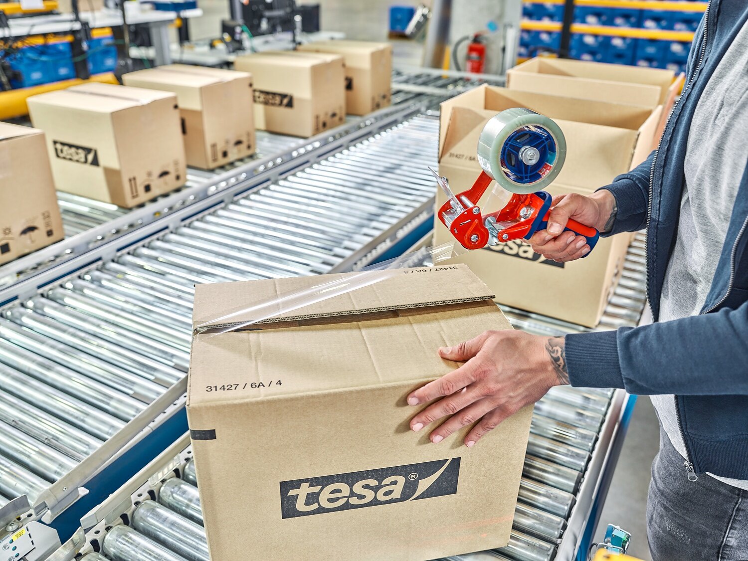 TESA-60412_MM_24744_final_72dpi A person seals a cardboard box labeled tesa on a conveyor belt in a warehouse using tesa tape. Several similar boxes can be seen in the background. The individual is using a red hand-held tesa tape dispenser. (This text has been generated by AI)