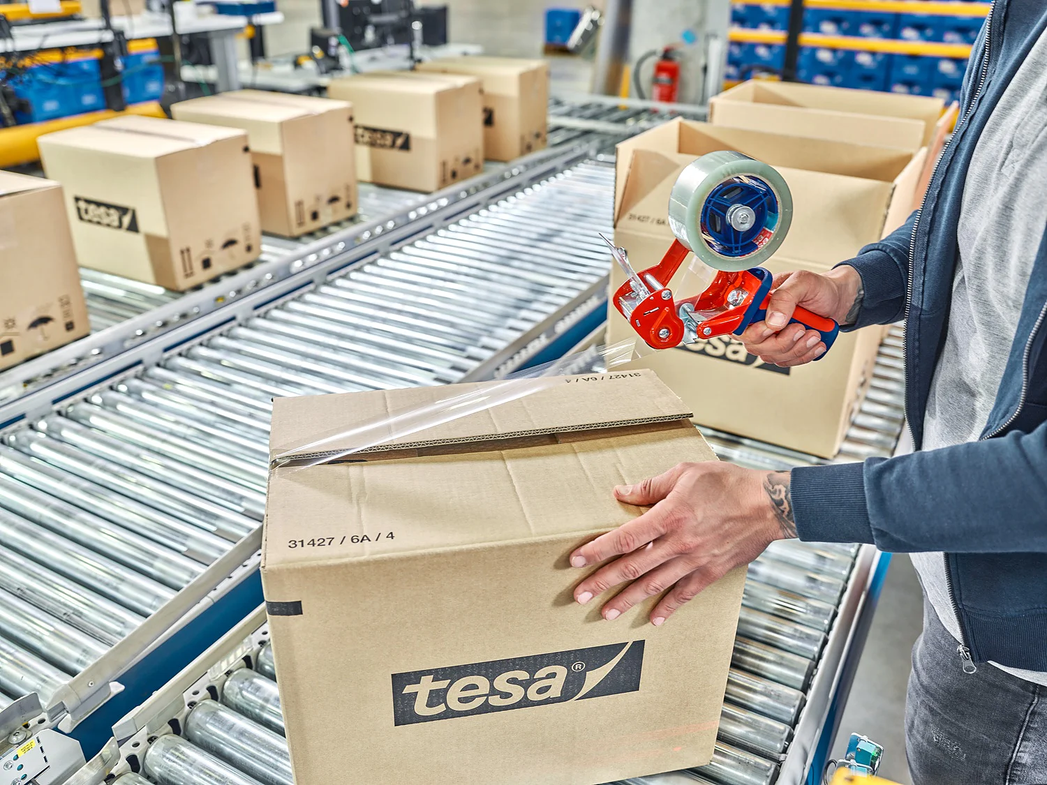 TESA-60412_MM_24744_final_72dpi A person seals a cardboard box labeled tesa on a conveyor belt in a warehouse using tesa tape. Several similar boxes can be seen in the background. The individual is using a red hand-held tesa tape dispenser. (This text has been generated by AI)