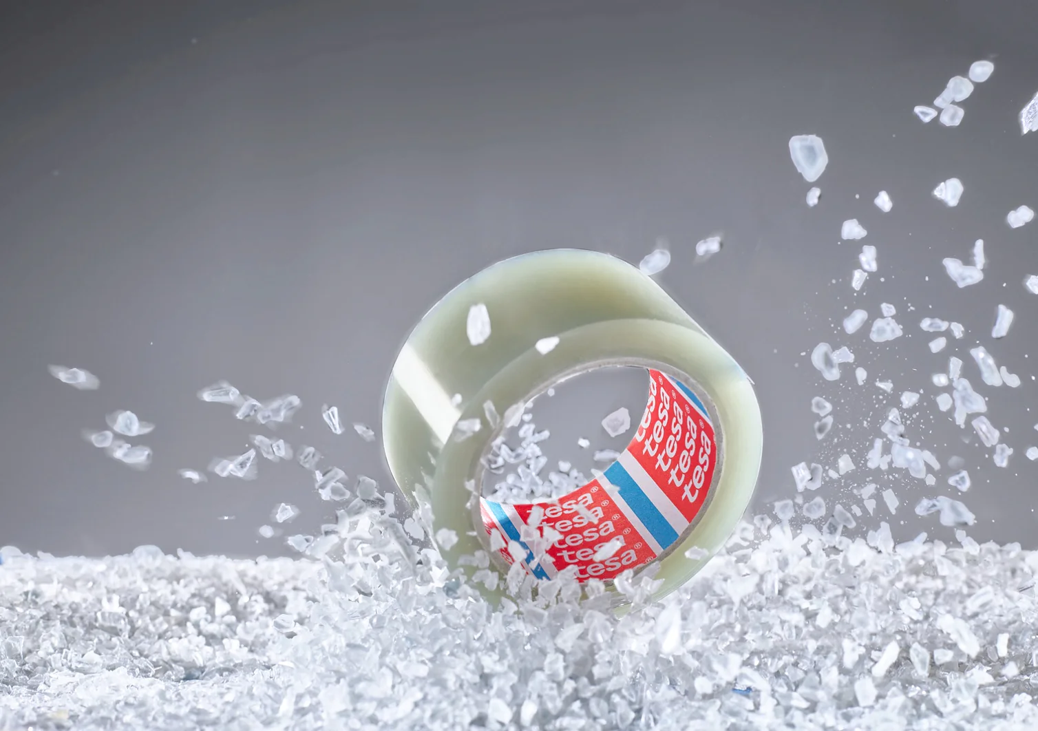 A roll of clear adhesive tesa tape with a red and white inner label floats amidst scattered translucent crystals against a gray background. The tesa tape is positioned upright, surrounded by the crystals in mid-air. (This text has been generated by AI)
