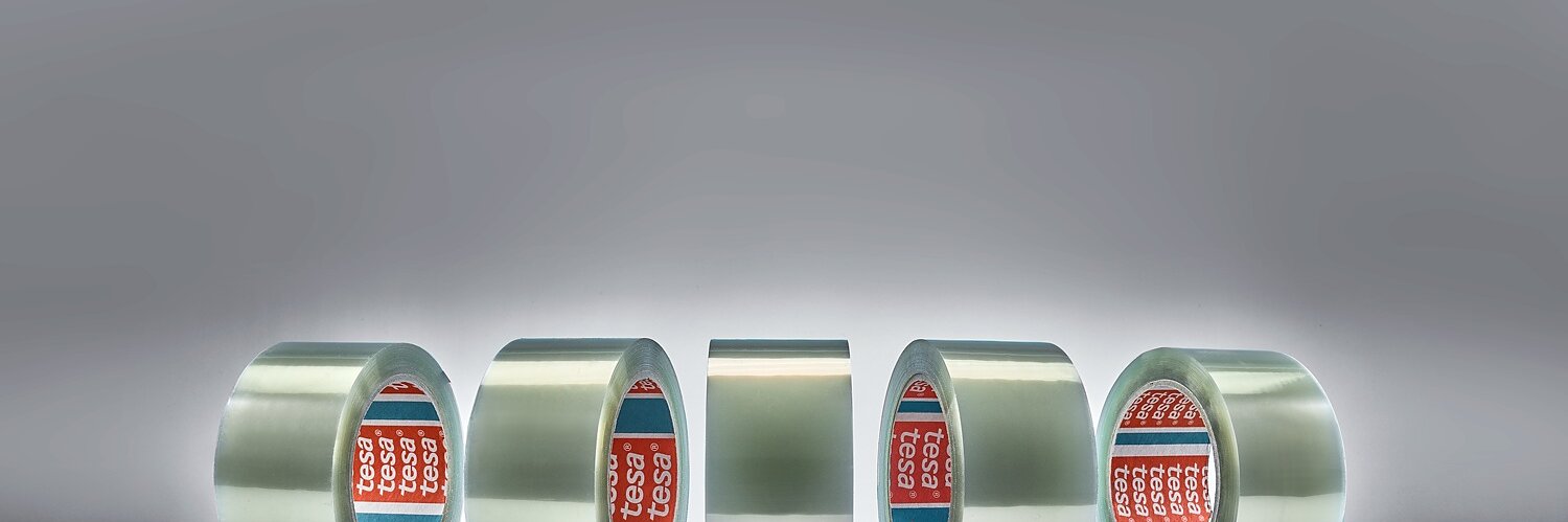 Five rolls of clear adhesive tesa tape are displayed vertically against a gray background. Small, scattered transparent granules are on the surface in front of the rolls. The tape cores are branded with red and blue labels.