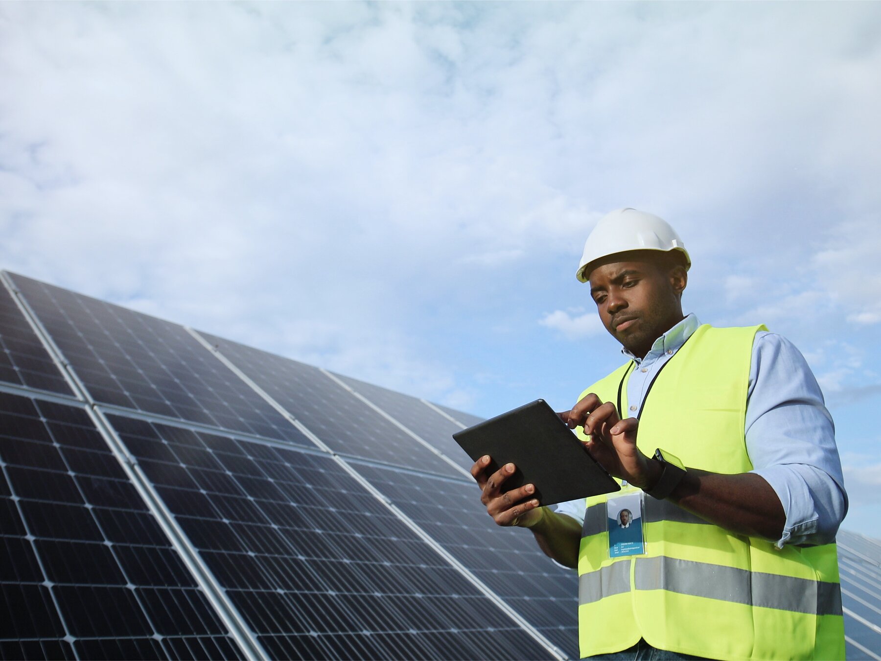 A person wearing a hard hat and neon safety vest is using a tablet near large solar panels secured with tesa tape. The sky is partly cloudy. (This text has been generated by AI)