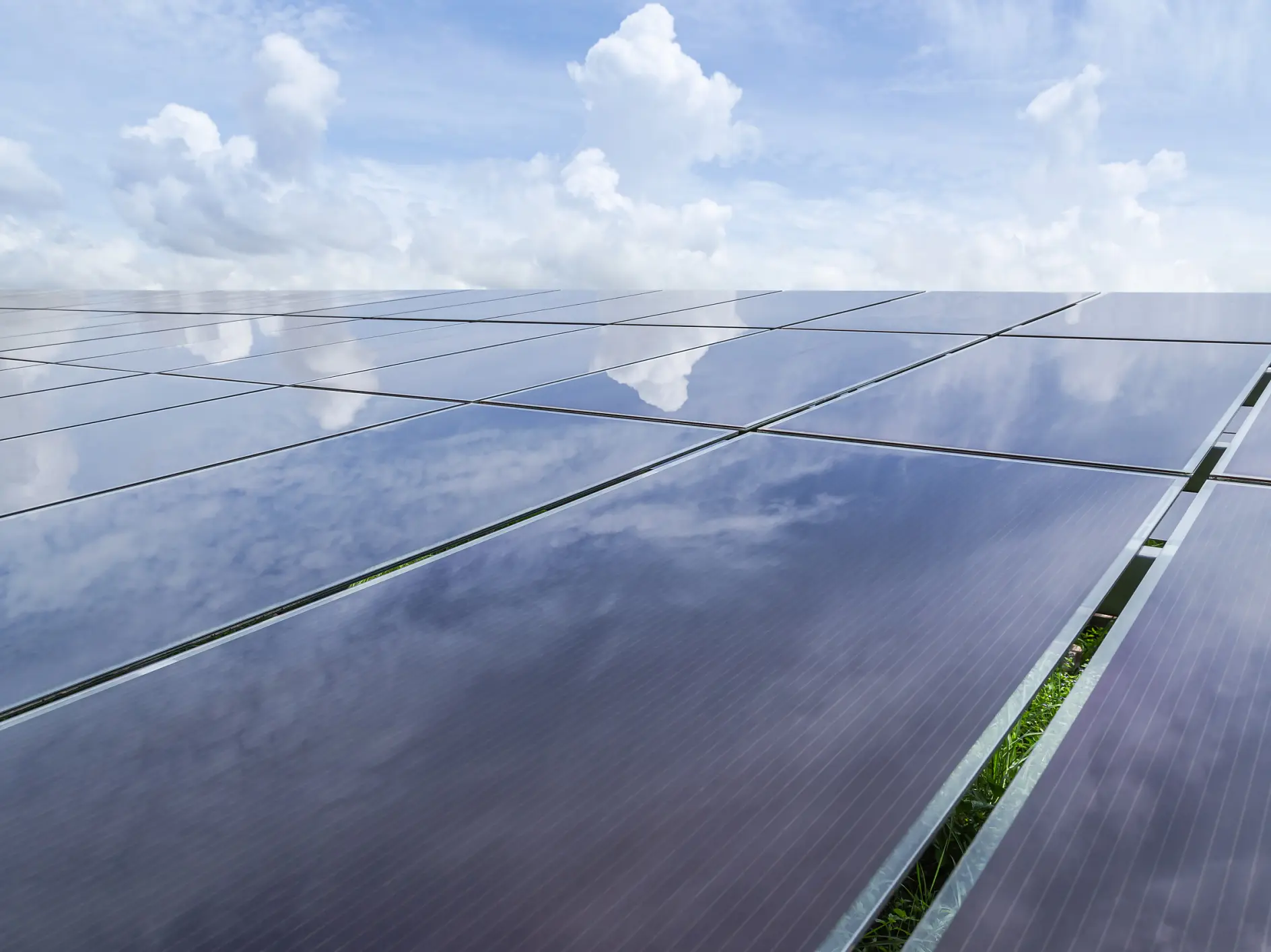 A large array of solar panels is seen reflecting a partly cloudy sky. The panels stretch across the image, capturing a mix of blue sky and white clouds. Vegetation is visible between the panels. (This text has been generated by AI)